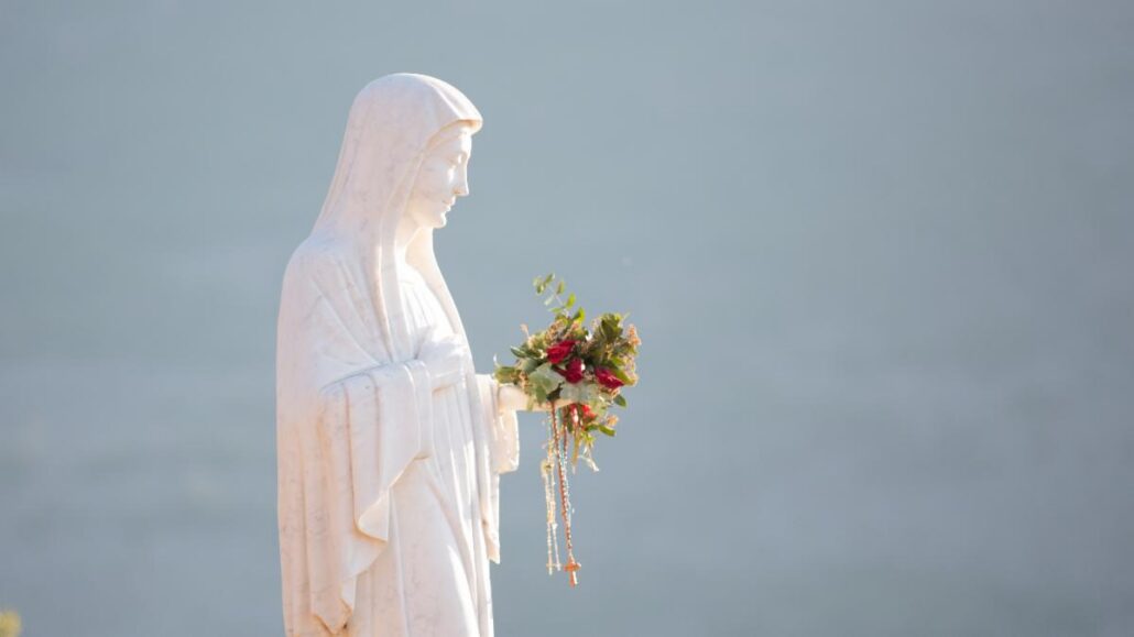 Statue of the Virgin Mary in white robes, holding a bouquet and rosary beads against a pale blue background.