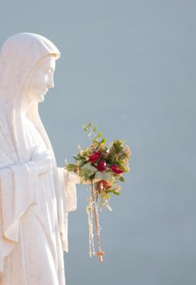 Statue of the Virgin Mary in white robes, holding a bouquet and rosary beads against a pale blue background.